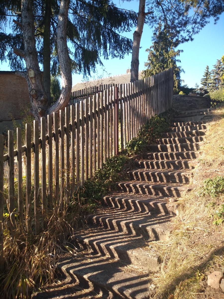 Na schody vedoucí nahoru vrhá stín laťkový plot a vytváří na schodech cik cak vedoucí čáry